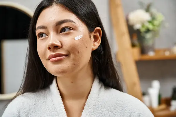 Crème de traitement immaculée sur le visage de la jeune femme asiatique avec l'acné regardant loin dans la salle de bain, soins de la peau — Photo de stock
