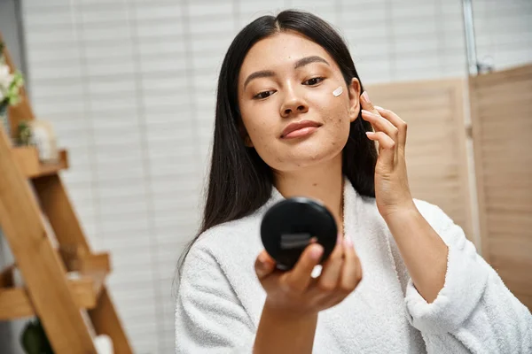 Crème de traitement imparfaite sur le visage de la jeune femme asiatique avec l'acné en regardant le miroir compact — Photo de stock