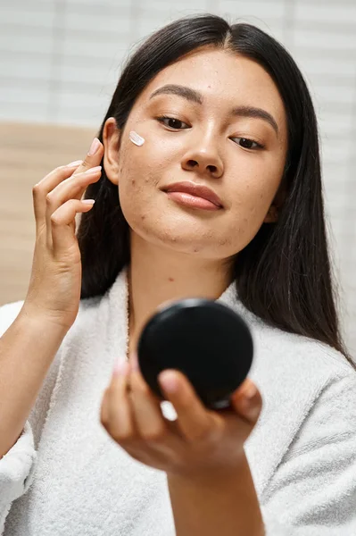 Blemish treatment cream on face of young asian woman with acne looking at compact mirror, vertical — Stock Photo
