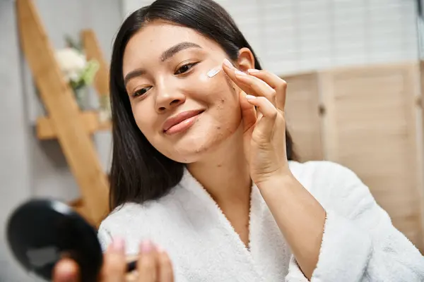 Jeune femme asiatique avec des cheveux bruns appliquant la crème de traitement de l'acné et en regardant le miroir compact — Photo de stock