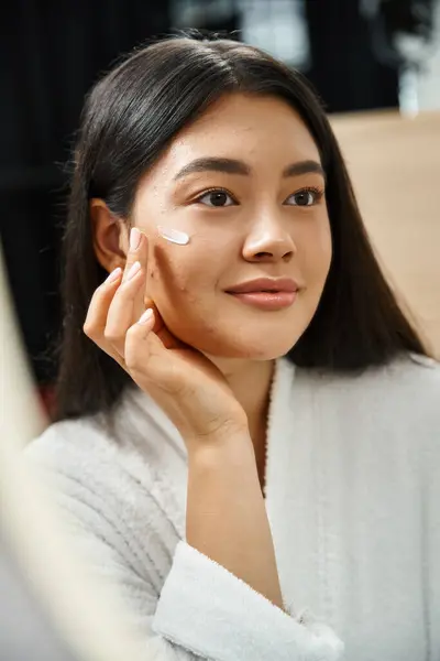 Фото азіатської жінки з брюнеткою волосся наносить крем для лікування акне і дивиться на дзеркало — стокове фото