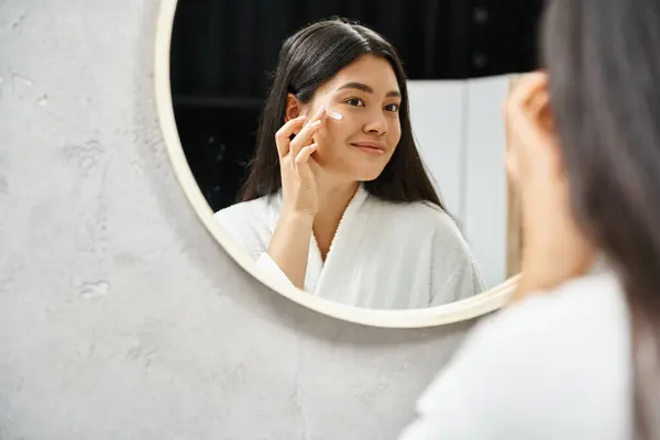 Alegre asiático mujer con morena cabello aplicando acné tratamiento crema y mirando espejo - foto de stock