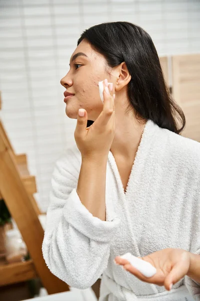 Side view of young asian woman with brunette hair and acne using facial cleansing foam in bathroom — Stock Photo