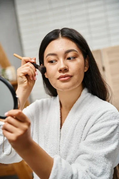 Mujer asiática joven con pelo morena aplicando maquillaje sobre la piel propensa al acné con cepillo cosmético - foto de stock