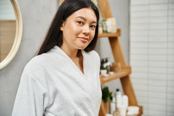 Portrait de jeune femme asiatique en peignoir debout dans la salle de bain moderne et regardant la caméra — Photo de stock
