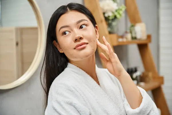 Retrato de jovem mulher asiática em roupão de banho tocando acne-propenso pele e olhando para longe no banheiro — Fotografia de Stock