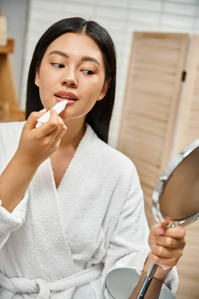 Young and brunette asian woman in bath robe applying lip balm and looking at mirror in bathroom — Stock Photo