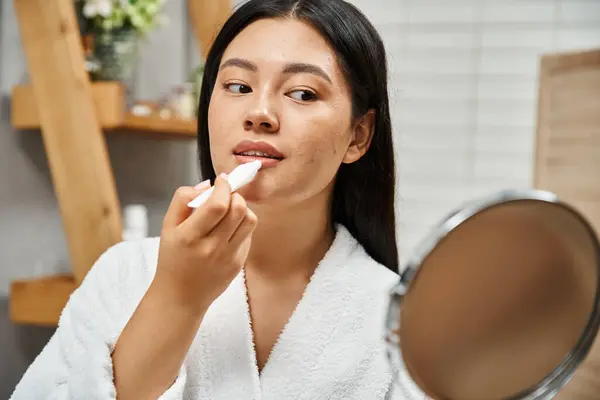 Morena joven asiática mujer en bata de baño aplicando bálsamo labial y mirando el espejo en el baño - foto de stock