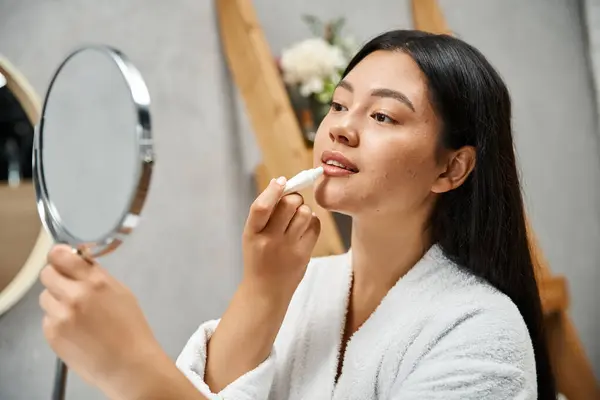 Joven y bonita mujer asiática en bata de baño aplicando bálsamo labial y mirando el espejo en el baño - foto de stock