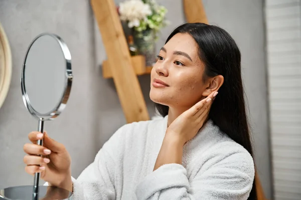 Brunette and young asian woman with acne-prone skin looking at mirror in modern bathroom, skin care — Stock Photo