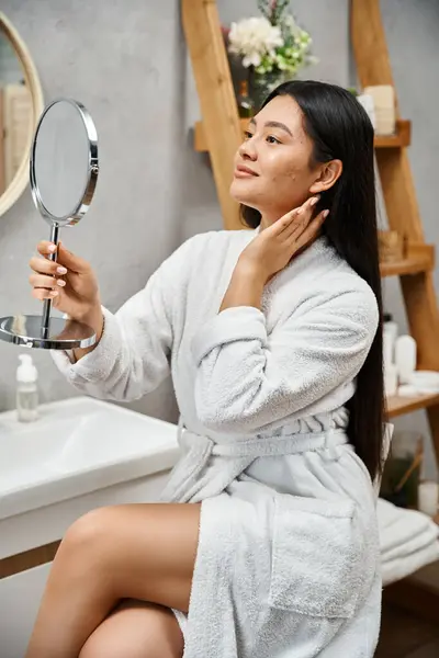 Ritratto di bruna e giovane donna asiatica con la pelle incline all'acne guardando lo specchio nel bagno moderno — Foto stock