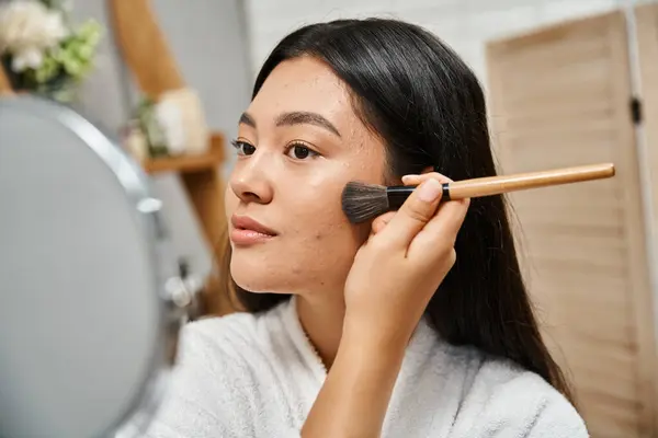 Молодая азиатская женщина с брюнетками и прыщами, нанося порошок на лицо и глядя на зеркало, проблемы с кожей — стоковое фото