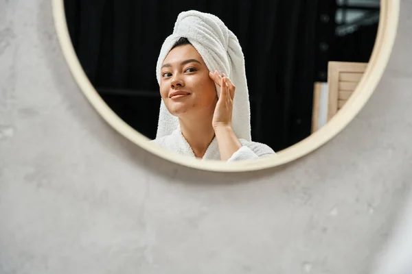 Jeune femme asiatique avec serviette blanche sur la tête toucher la peau sujette à l'acné et en regardant miroir de salle de bains — Photo de stock