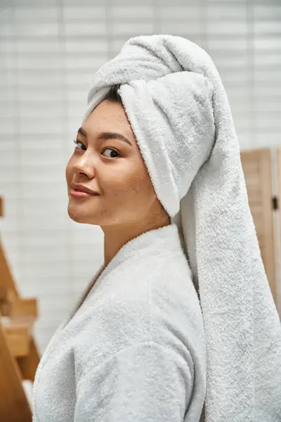 Heureux et jeune asiatique femme en robe avec serviette blanche sur la tête en regardant caméra dans salle de bain — Photo de stock
