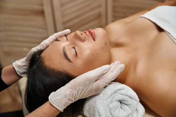 Massage facial pour la peau sujette à l'acné, thérapeute en gants de latex et client asiatique avec les yeux fermés — Photo de stock