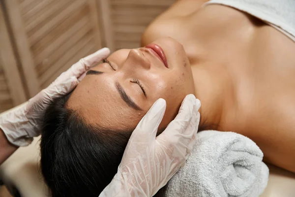 Terapeuta em luvas de látex fazendo massagem facial para asiático cliente com olhos fechados e acne propenso a pele — Fotografia de Stock
