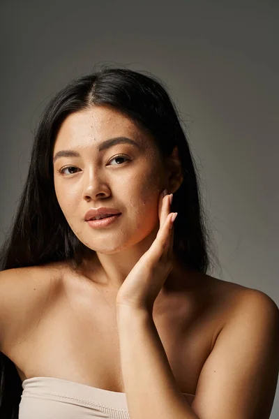 Vertical view of young asian woman with skin issues and bare shoulders looking at camera on grey — Stock Photo