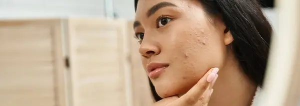 Junge asiatische Frau mit brünetten Haaren und zu Akne neigender Haut beim Anblick des Spiegels im Badezimmer, Banner — Stockfoto
