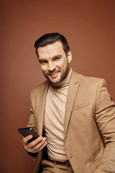 Стильний і красивий чоловік з щетиною посміхається і тримає смартфон на бежевому тлі, соціальні медіа — стокове фото