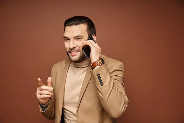 Handsome man in stylish blazer smiling while having phone call on smartphone, beige backdrop — Stock Photo