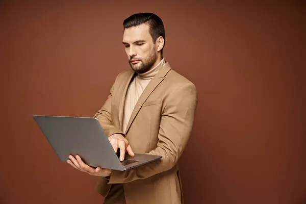 Determinado y guapo profesional utilizando su ordenador portátil mientras trabaja de forma remota sobre fondo beige - foto de stock