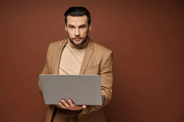 Focalizzato e bello professionista che tiene il suo computer portatile mentre lavora a distanza su sfondo beige — Foto stock