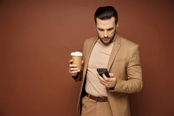 Uomo in abbigliamento elegante che tiene il caffè per andare e utilizzando il suo smartphone su sfondo beige, digitale — Foto stock