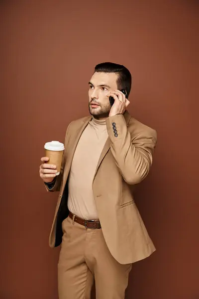 Handsome man in stylish attire holding coffee to go and having call on smartphone on beige backdrop — Stock Photo