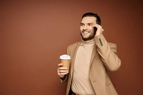 Homme heureux en tenue élégante tenant café pour aller et ayant appel sur smartphone sur fond beige — Photo de stock