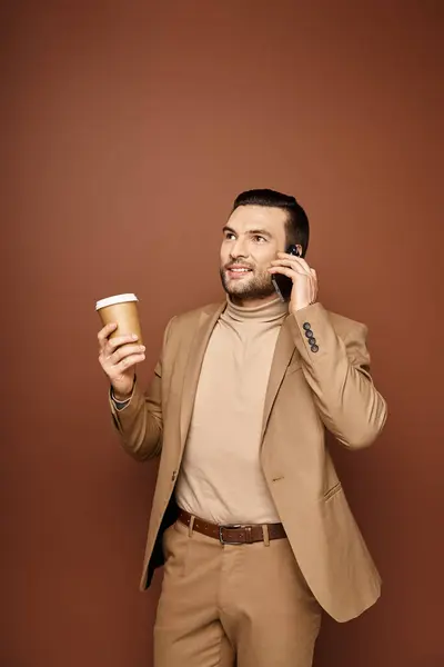 Content man in stylish attire holding coffee to go and having call on smartphone on beige backdrop — Stock Photo