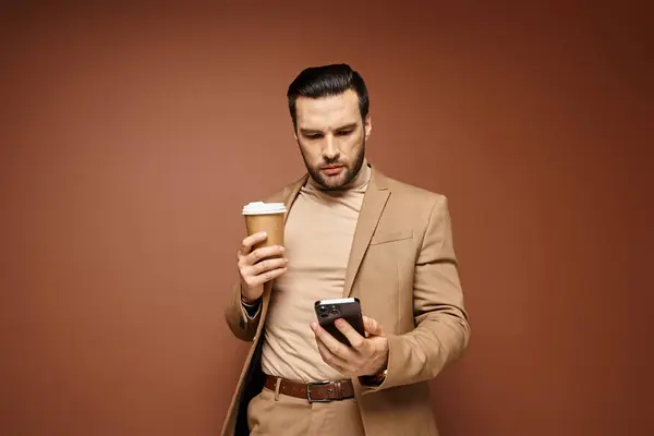 Bell'uomo in abbigliamento elegante che tiene il caffè per andare e usando il suo smartphone su sfondo beige — Foto stock