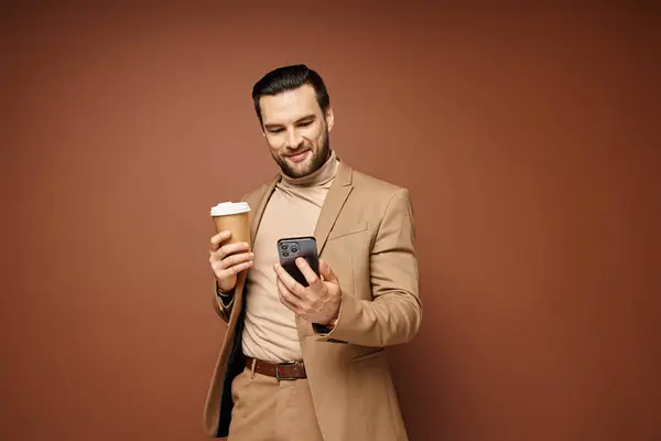 Happy man in stylish attire holding coffee to go and using his smartphone on beige background — Stock Photo