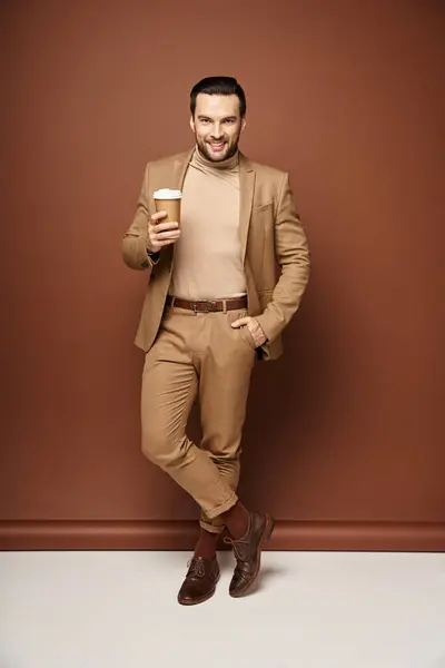 Zufriedener Mann in stylischer Kleidung mit Coffee to go und posiert mit der Hand in der Tasche vor beigem Hintergrund — Stockfoto