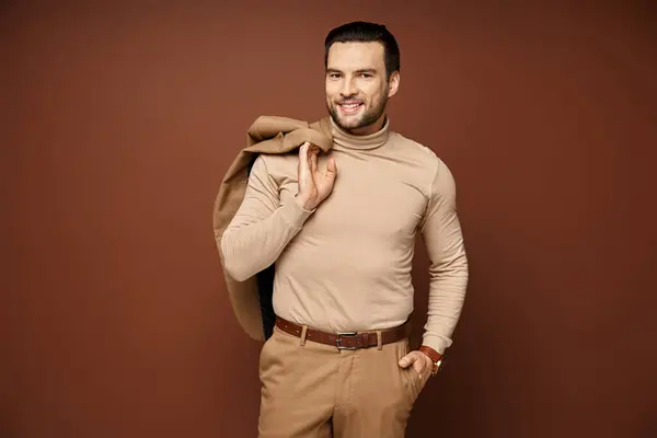 Happy man in turtleneck holding his jacket over shoulder and posing with hand in pocket on beige — Stock Photo