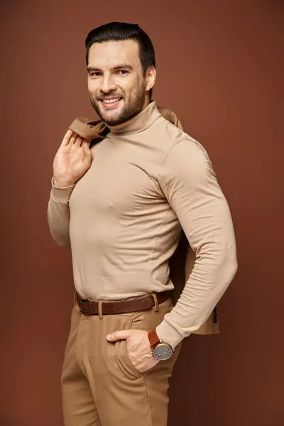 Hombre feliz con cuello alto sosteniendo chaqueta de moda sobre el hombro y posando con la mano en el bolsillo en beige - foto de stock
