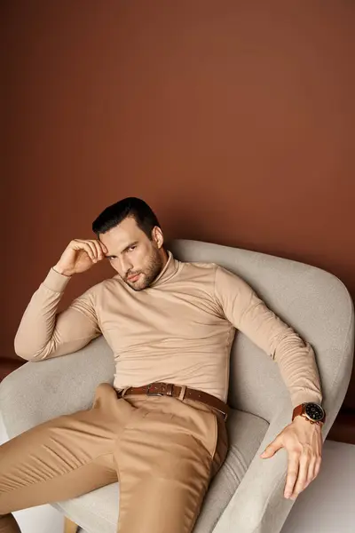 Pensive man in turtleneck sitting on comfortable armchair and looking at camera on beige backdrop — Stock Photo