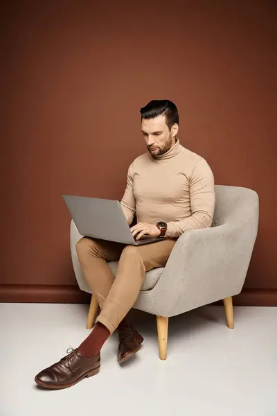 Красивий чоловік в черепашці сидить на зручному кріслі і використовує ноутбук на бежевому тлі — стокове фото