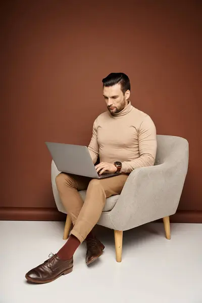 Bell'uomo in dolcevita seduto su una comoda poltrona e utilizzando il computer portatile su sfondo beige — Foto stock
