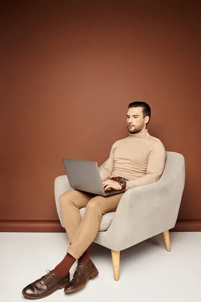 Hombre de buen aspecto en cuello alto sentado en un sillón cómodo y el uso de la computadora portátil en el fondo beige - foto de stock