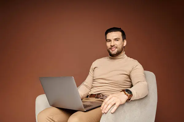 Cheerful man in turtleneck sitting on comfortable armchair and using laptop on beige backdrop — Stock Photo