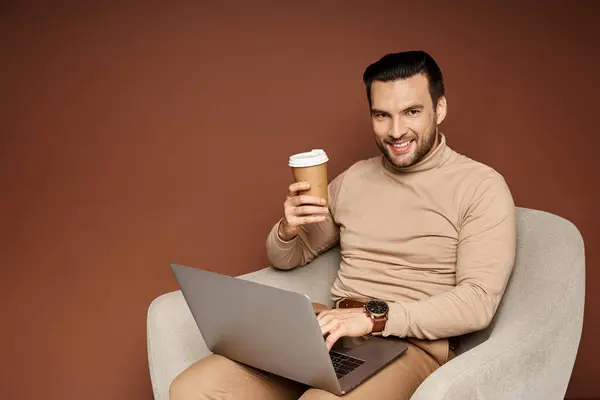 Cheerful man using laptop and holding coffee to go while sitting on armchair on beige backdrop — Stock Photo