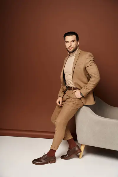Gut aussehender Mann im stylischen Anzug posiert mit der Hand in der Tasche in der Nähe eines Sessels auf beigem Hintergrund — Stockfoto
