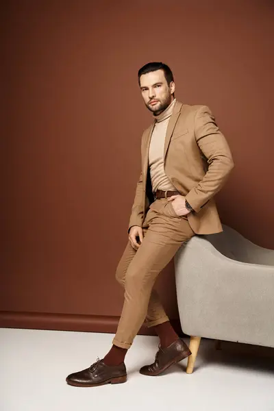 Schöner Mann im eleganten Anzug posiert mit der Hand in der Tasche in der Nähe eines Sessels auf beigem Hintergrund — Stockfoto