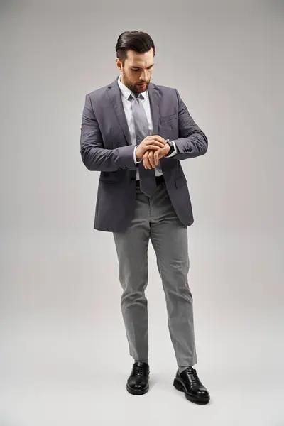 Handsome businessman posing in a sleek suit checking his wristwatch on grey background, elegance — Stock Photo