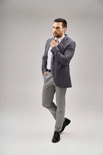 Schöner Mann in scharfem und stilvollem Anzug, der seine Krawatte anpasst und mit der Hand in der Tasche auf grau posiert — Stockfoto