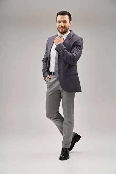 Homem feliz em terno elegante ajustando sua gravata e posando com a mão no bolso no fundo cinza — Fotografia de Stock