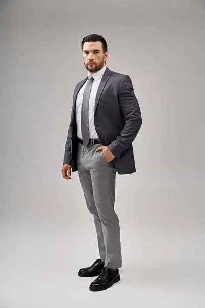 Hombre guapo en traje afilado y elegante posando con la mano en el bolsillo sobre fondo gris, elegancia - foto de stock