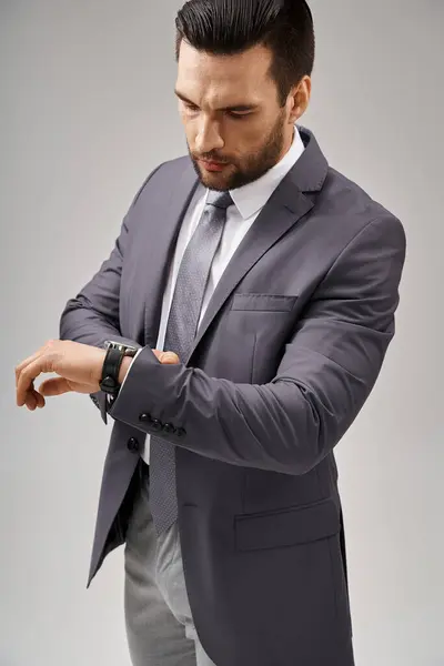 Hombre de negocios guapo posando en un traje elegante revisando su reloj de pulsera sobre fondo gris, elegancia - foto de stock