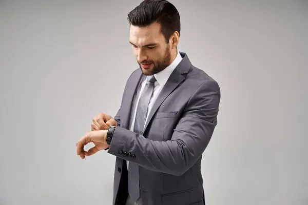 Homem de negócios bonito posando em terno elegante verificando seu relógio de pulso em fundo cinza, pontual — Fotografia de Stock