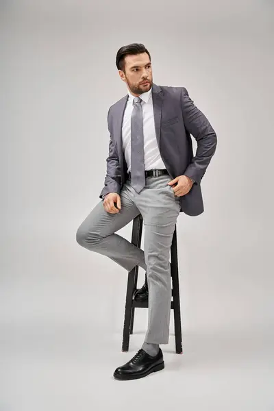 Uomo d'affari bello e sicuro di sé in abbigliamento formale appoggiato su sgabello alto su sfondo grigio — Foto stock